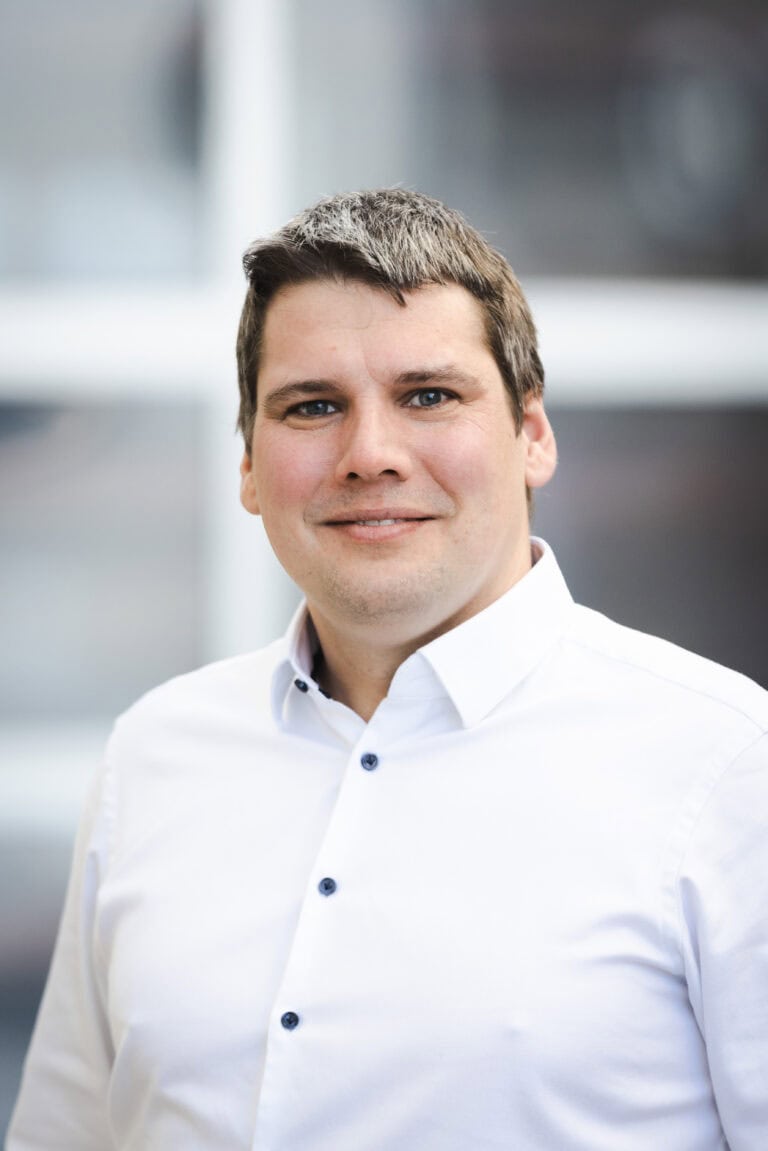 A professional portrait of a smiling man in a white shirt, standing outdoors with a blurred modern building background, representing corporate leadership and confidence.