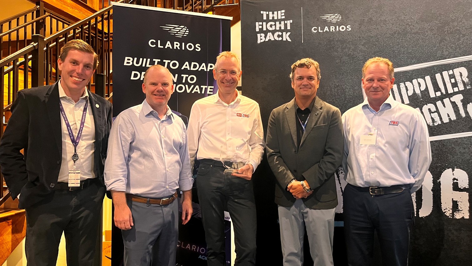 Group of five men standing together at a professional event, with banners displaying 'Clarios' and 'The Fight Back' in the background, representing Rosendahl Nextrom's industry presence.