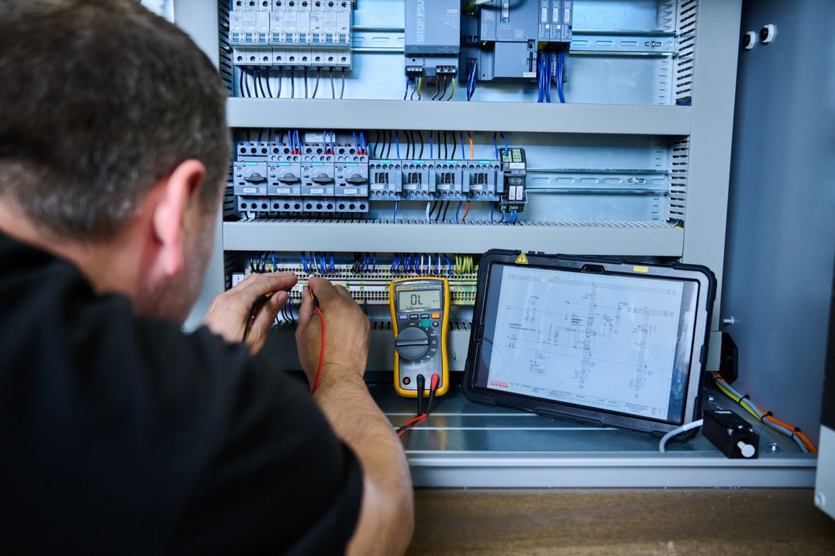 An electrician testing electrical control panel with a multimeter and tablet for automation system diagnostics at Rosendahl Nextrom facility.