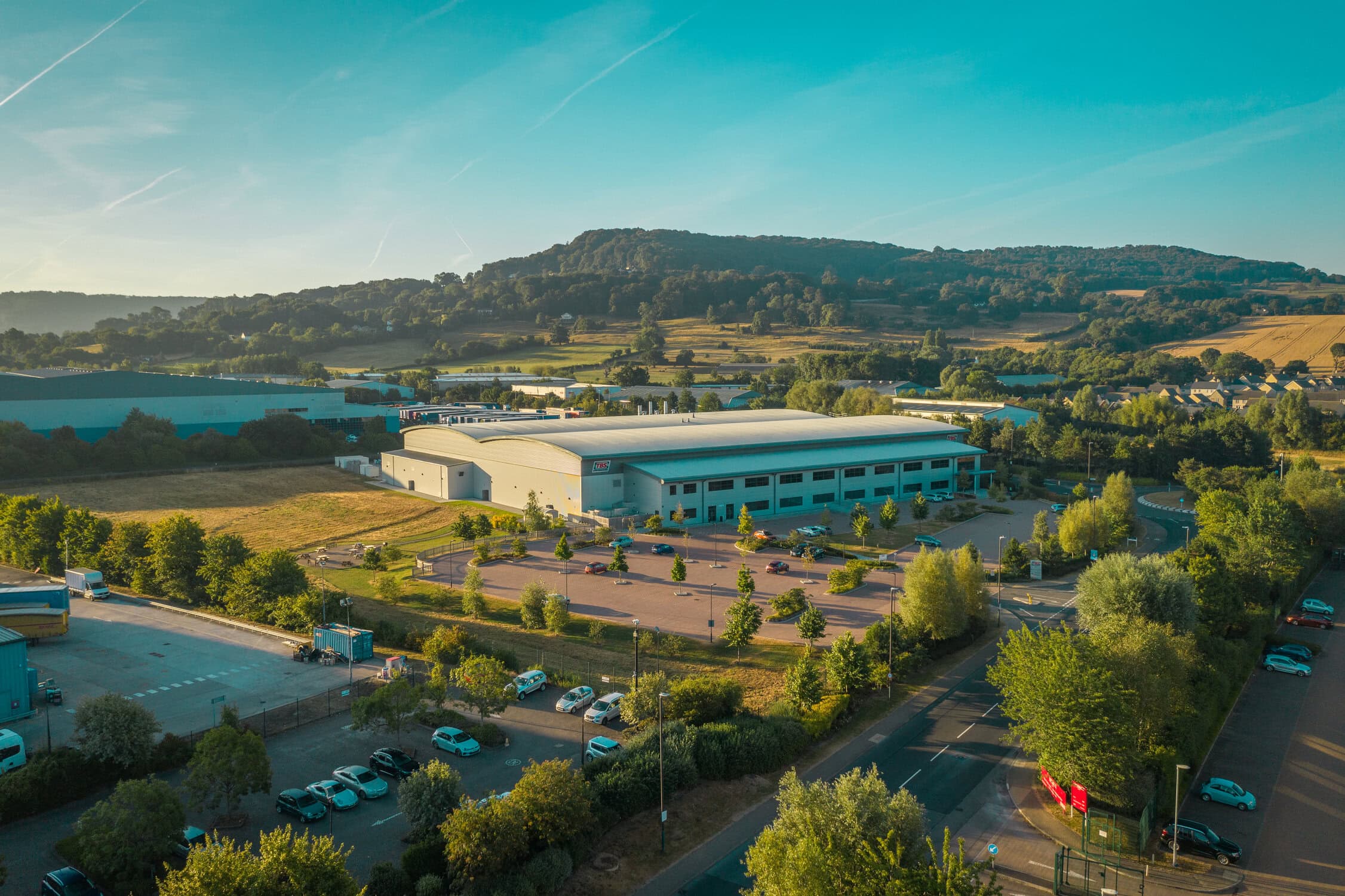Aerial view of Rosendahl Nextrom manufacturing facility surrounded by greenery and parking lots, with rolling hills in the background under a clear blue sky.