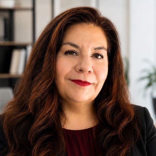 A professional woman with long, wavy auburn hair, wearing a black blazer and red top, in a modern office setting with bookshelves and plants, representing business and corporate environment.